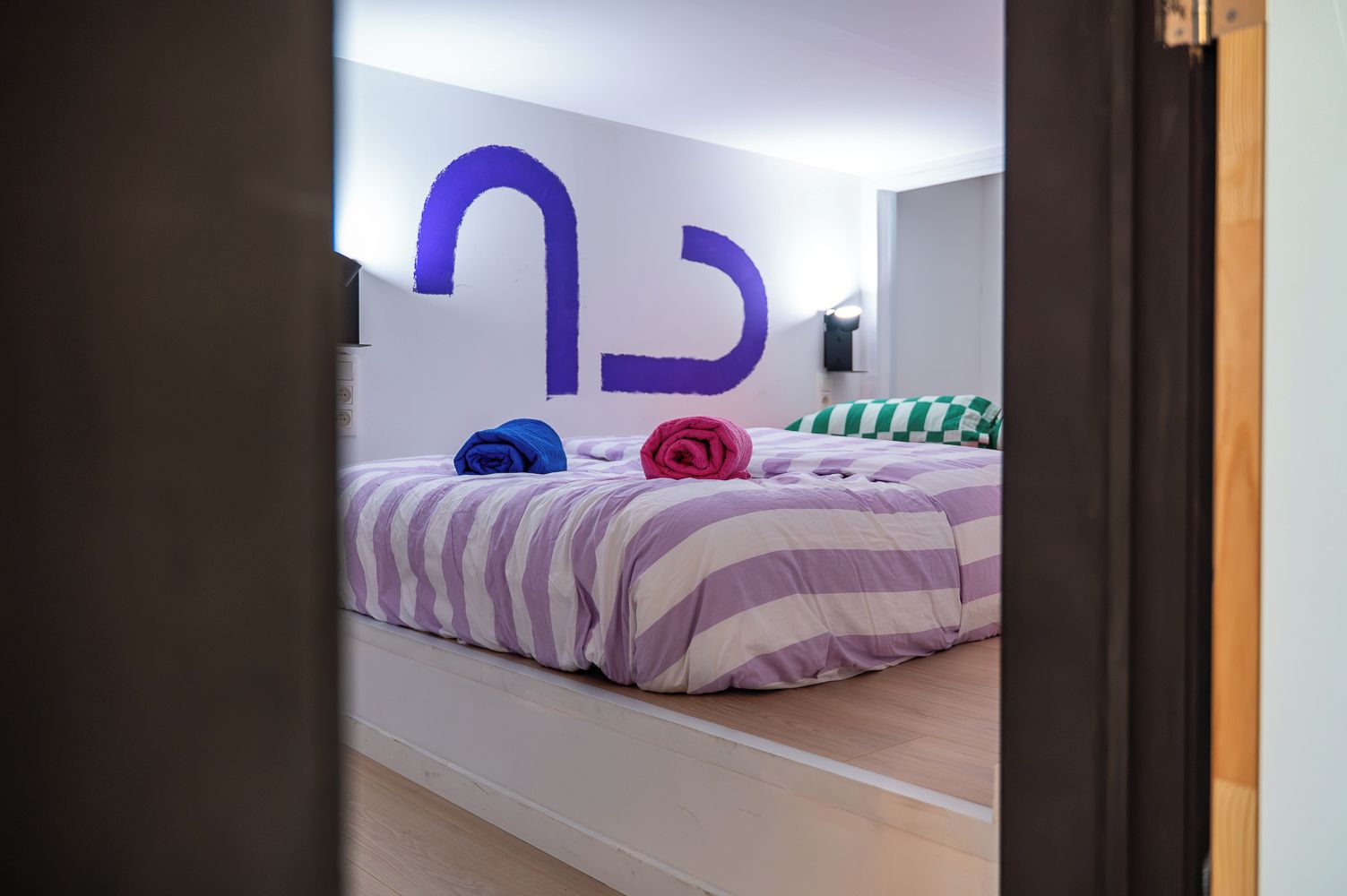 Une pièce avec un lit, une grande lettre violette peinte sur le mur et un cadre de porte.