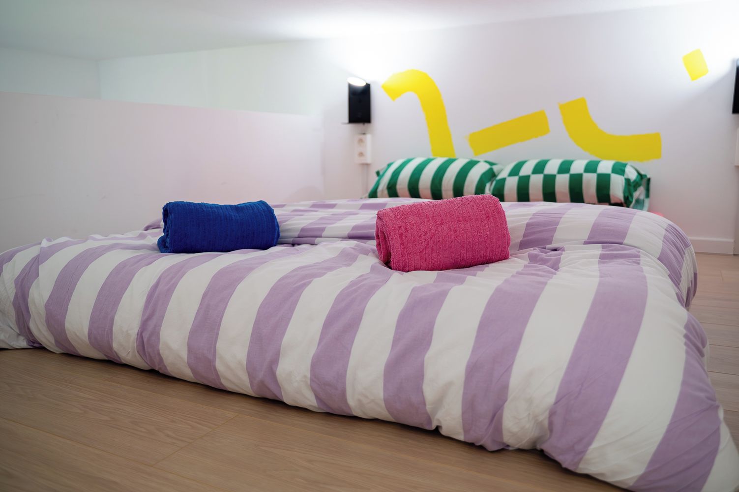 Een bed met gestreepte lakens en twee opgerolde handdoeken, een wand met een abstract geel ontwerp, en een houten vloer.