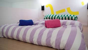 Een bed met gestreepte lakens en twee opgerolde handdoeken, een wand met een abstract geel ontwerp, en een houten vloer.
