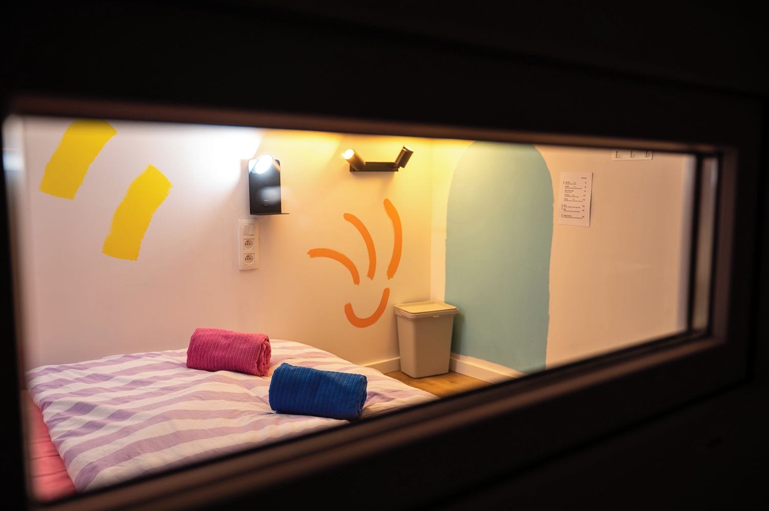 Et værelse med en seng, en skraldespand og en lampe på væggen.