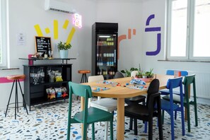 Ein modernes Interieur mit einem Holztisch, bunten Stühlen, einem schwarzen Bücherregal und einem Neon-Leuchtschild.