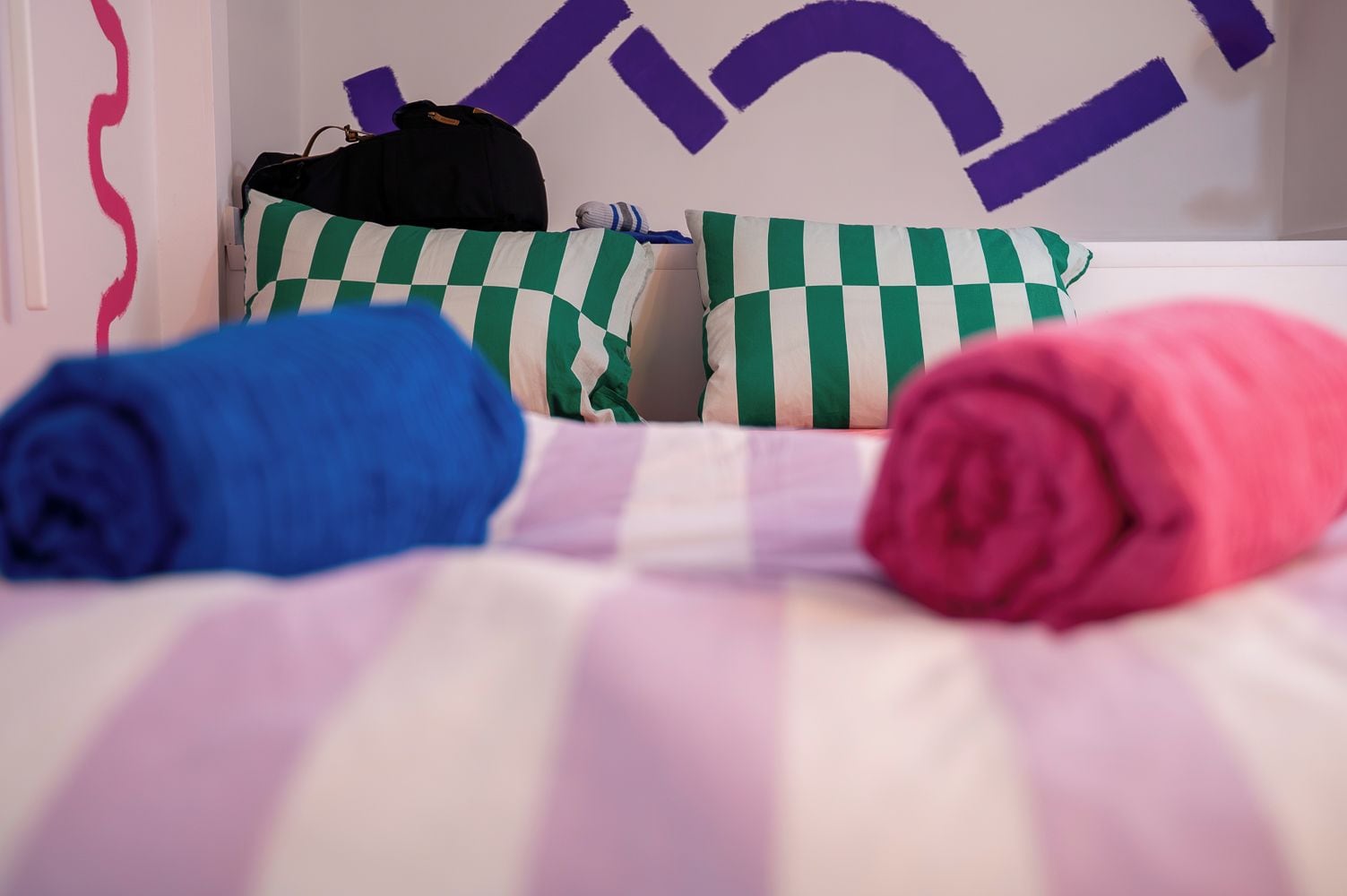 Una cama con ropa de cama a rayas, una almohada verde y blanca a cuadros y toallas enrolladas en azul y rosa.