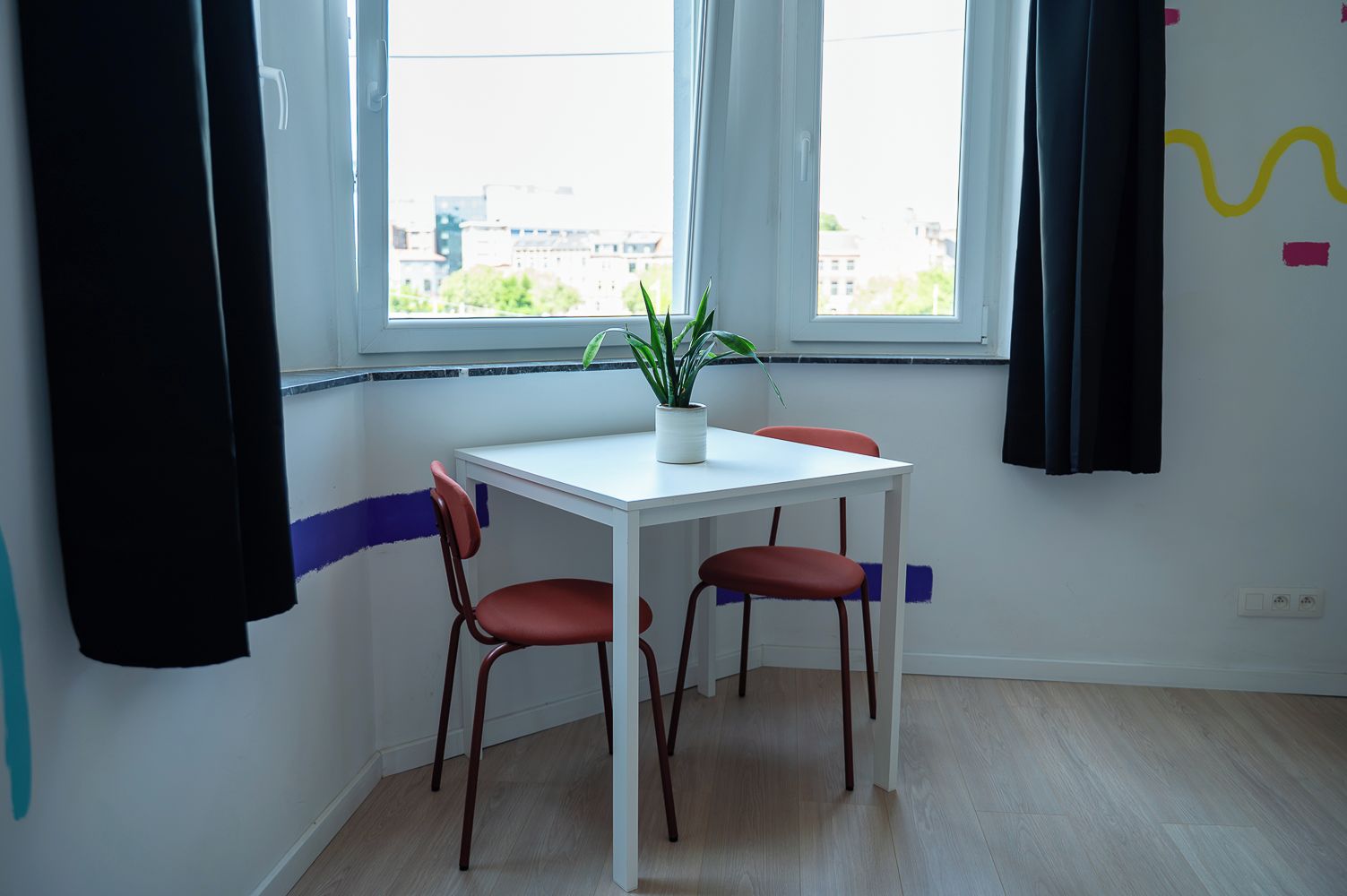 Une pièce avec une table blanche, deux chaises rouges, une plante en pot et une fenêtre avec des rideaux.