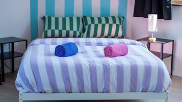 Una cama con ropa de cama a rayas, dos toallas enrolladas, una mesita de noche con una lámpara y un cojín de cuadros.