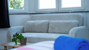Een kamer met een grijze bank, een bijzettafeltje met een plant en een raam met gordijnen.