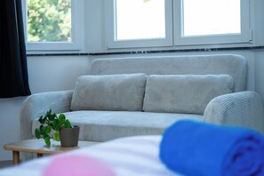 Ein Zimmer mit einem grauen Sofa, einem kleinen Tisch mit Pflanze und einem Fenster mit Vorhängen.