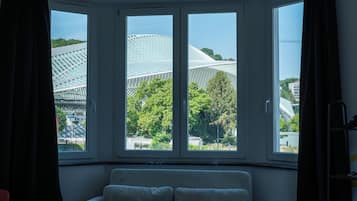 Une pièce dotée d’une baie vitrée donnant sur un bâtiment moderne à la façade en verre, entouré d’arbres.