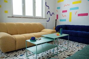 A room with a yellow sofa, a blue sofa, a glass coffee table, and a patterned floor. - Cool Cool House (Liège)