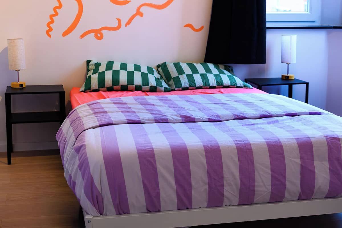 A bed with a purple and white striped bedspread, green and white checkered pillows, and a red blanket. There are two bedside tables with lamps, one on each side of the bed.