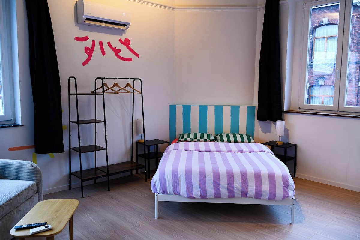 A bedroom with a bed, a nightstand, a lamp, a shelf unit, and a window.