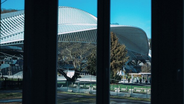 A modern building with a curved roof structure, surrounded by a fenced area with trees and a clear sky. - Cool Cool House (Liège)