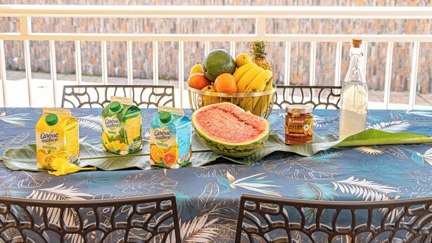 A table with a tropical tablecloth, a fruit basket, juice cartons, a watermelon, and a bottle of honey. - Quiet 3-suite house with 4 bedrooms - Assured water - Between sea and mountains. (Baie-Mahault)
