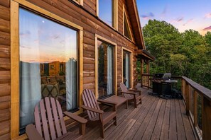 Une terrasse en bois avec des fauteuils Adirondack, un barbecue et une vue sur l’intérieur d’un chalet.