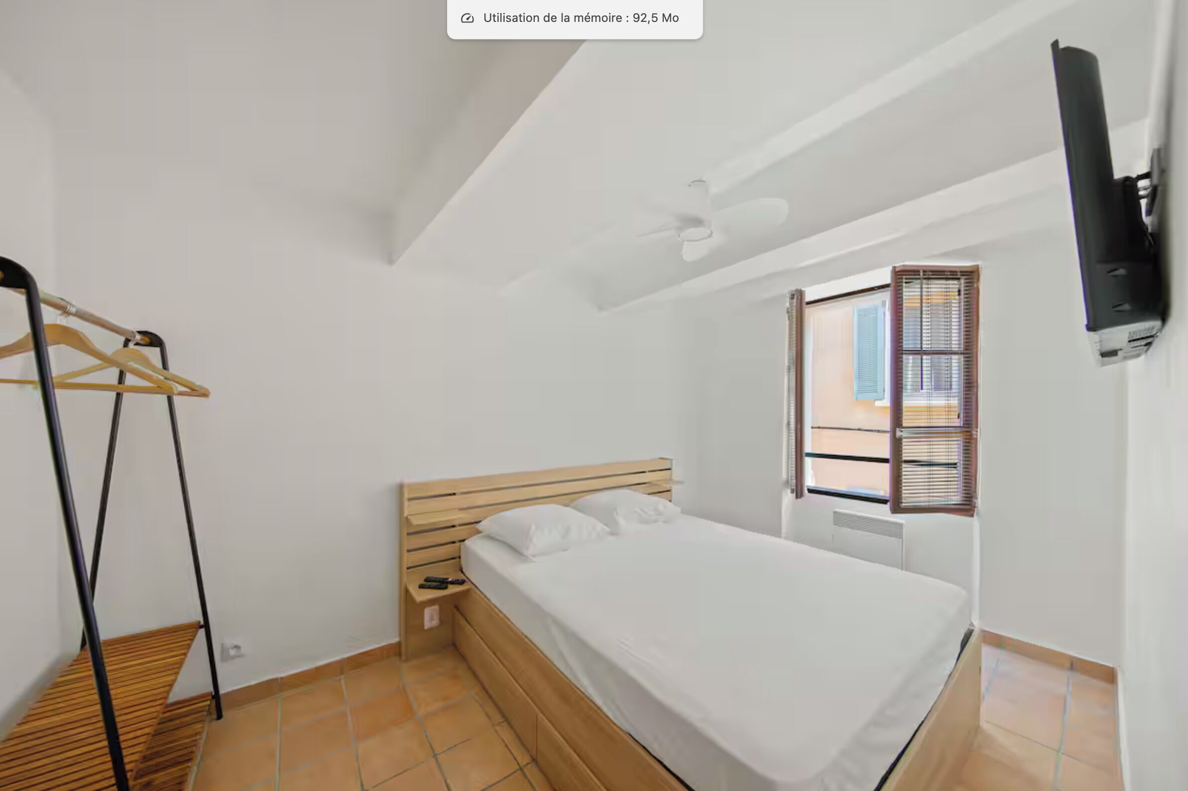 Une chambre à coucher avec un lit, une tête de lit en bois, un ventilateur de plafond, un téléviseur fixé au mur et une fenêtre avec des stores.