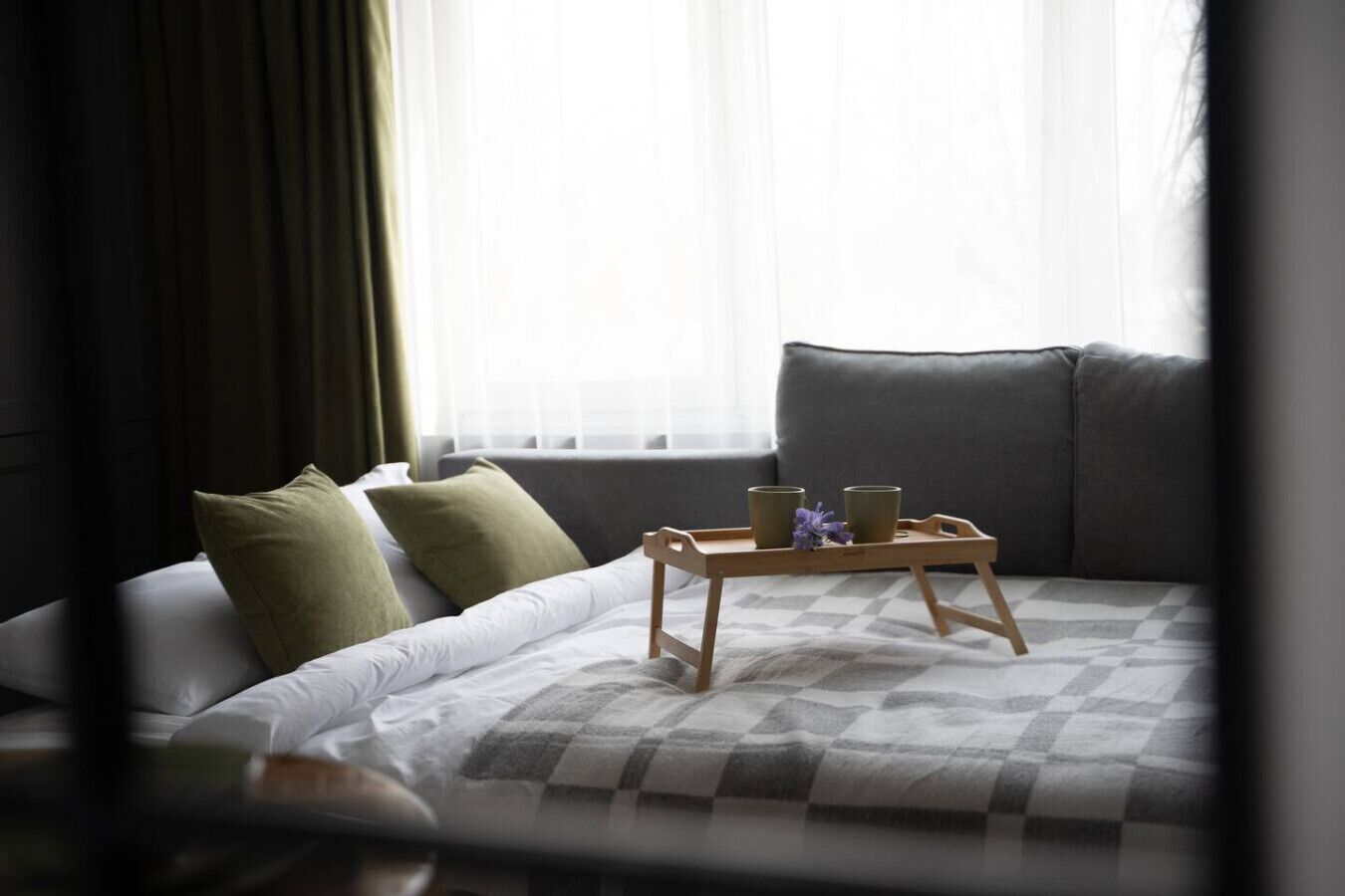 A neatly made bed with a checkered blanket, a wooden tray with two cups and a flower, and a grey sofa in a room with sheer curtains.
