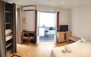 A hotel room with a bed, a desk with a chair, a TV, and a kitchenette. - Cozy 2-Person Private Parking Spot - Near the Beach & Parks (Antibes)