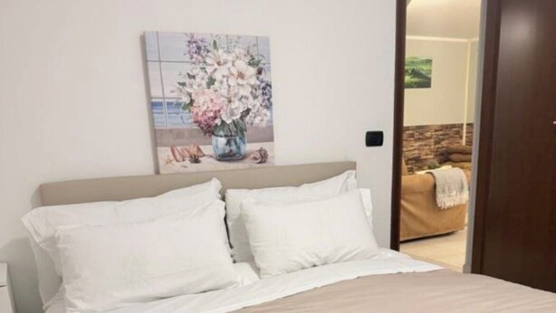 Una cama bien hecha con sábanas blancas y una manta de cuadros, un cuadro de flores en la pared, y vista de una sala a través de una puerta abierta.