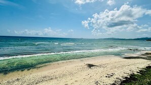 Ein Strand mit klarem Wasser, sandigem Ufer und fernen Bergen unter einem teils bewölkten Himmel.