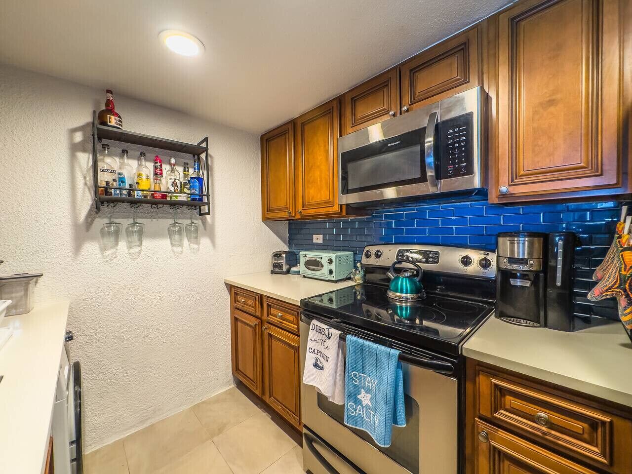 A kitchen with wooden cabinets, a stainless steel microwave, a black stove, a coffee maker, and a shelf with bottles and glasses.