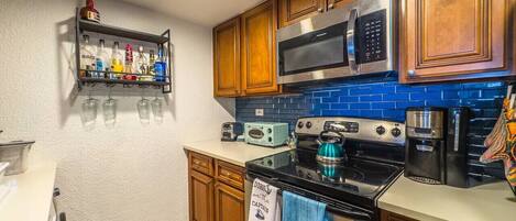 A kitchen with wooden cabinets, a stainless steel microwave, a black stove, a coffee maker, and a shelf with bottles and glasses.