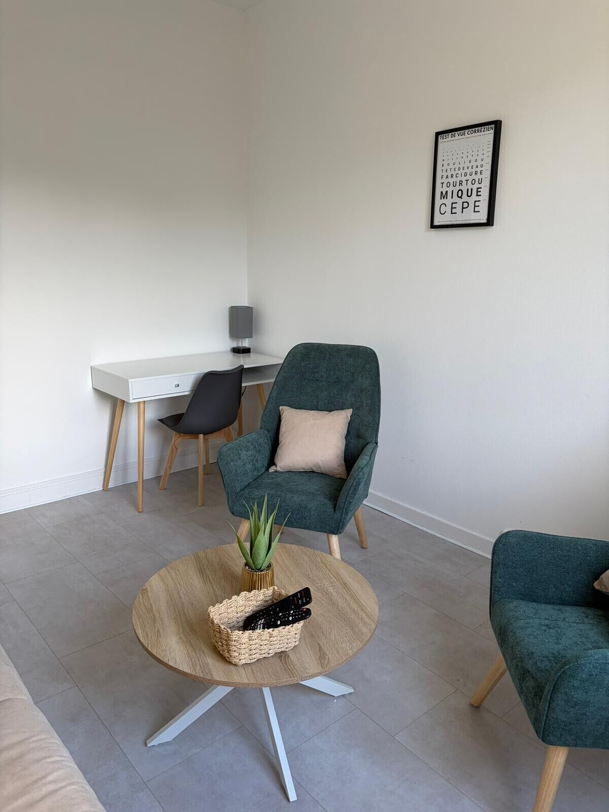 Ein modernes Wohnzimmer mit einem petrolfarbenen Sessel, einem runden Couchtisch mit Pflanze und Fernbedienungen, einem weißen Schreibtisch mit Lampe und einer an der Wand befestigten Sehschublade.