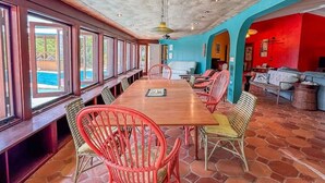 A spacious dining area with a long table and multiple chairs, large windows along one wall, and a vibrant interior with a red and blue color scheme. - Panorama Pool House - 3 BR/3BA! (Christiansted)
