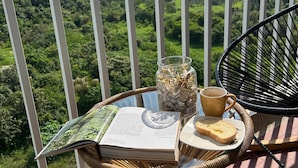 Ein Korbtisch mit einem Buch, einem Glas, einer Tasse und einem Teller mit Toast auf einem Balkon mit Blick auf eine Berglandschaft.