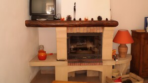 Une cheminée surmontée d’une étagère en bois où est posée une télévision, une petite table avec une bougie et des pommes de pin, et une lampe sur la droite.