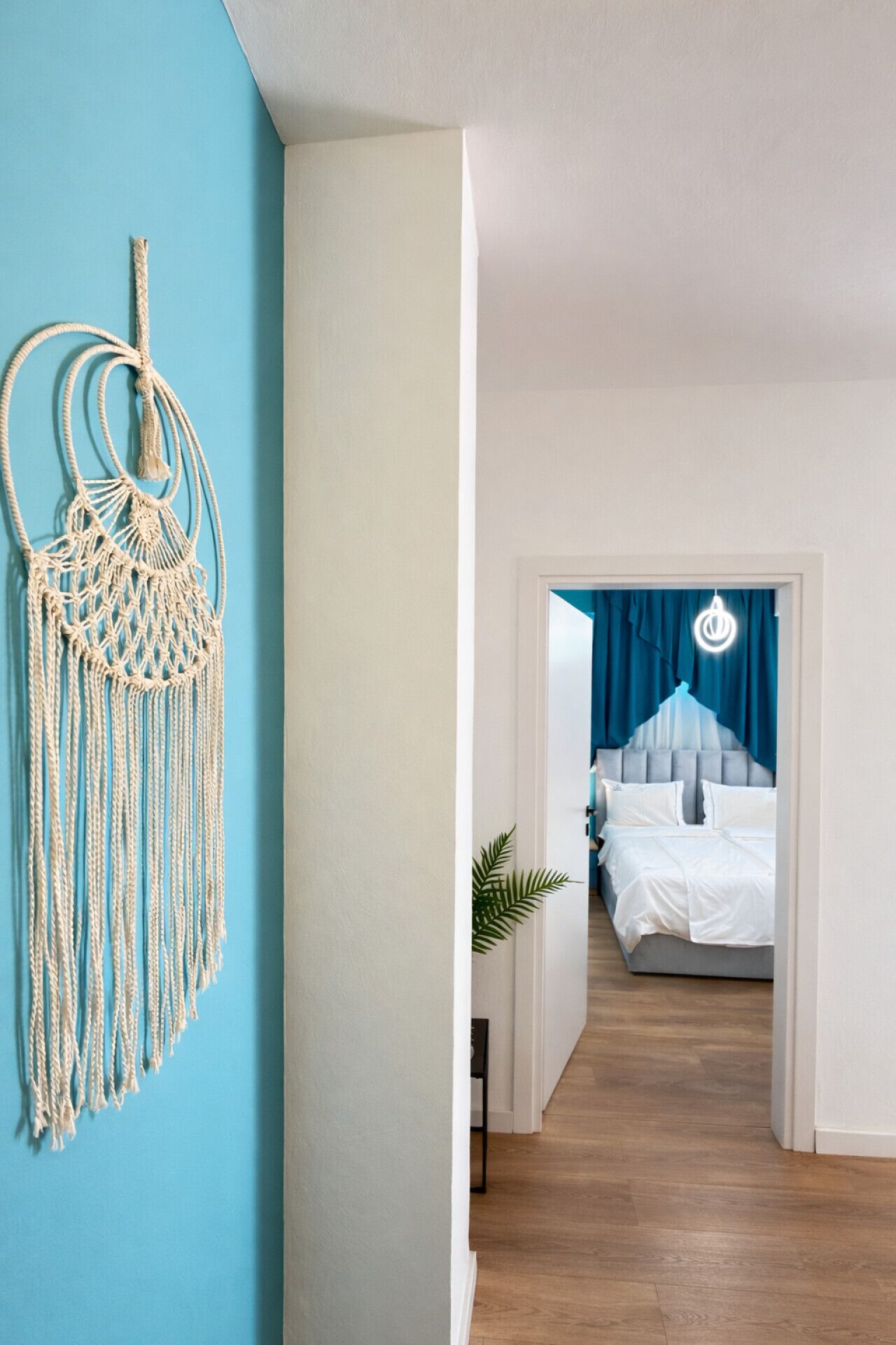 Un couloir menant à une chambre à coucher, où se trouvent un lit, une décoration murale en macramé et une plante.