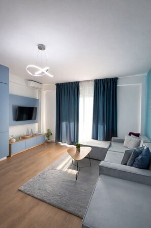 A modern living room with a grey sofa, a coffee table, and a TV mounted on the wall. There is a window with blue curtains, a ceiling light fixture, and a small plant on the floor. - Tiffany`s Apartment Sea View (Durrës)