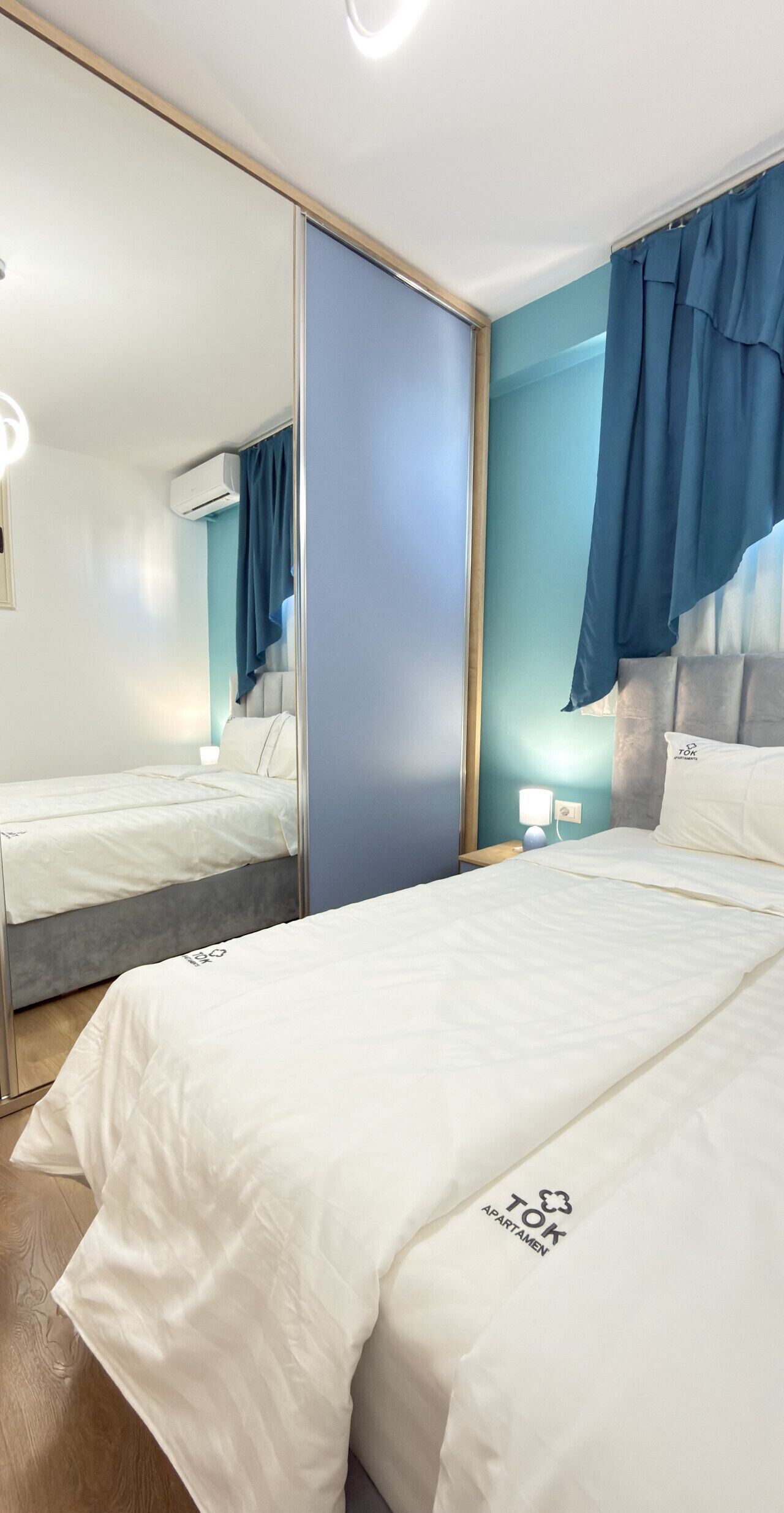 Een hotelkamer met een grote spiegel, een bed met witte lakens, een nachtkastje met een lamp en een blauw gordijn.