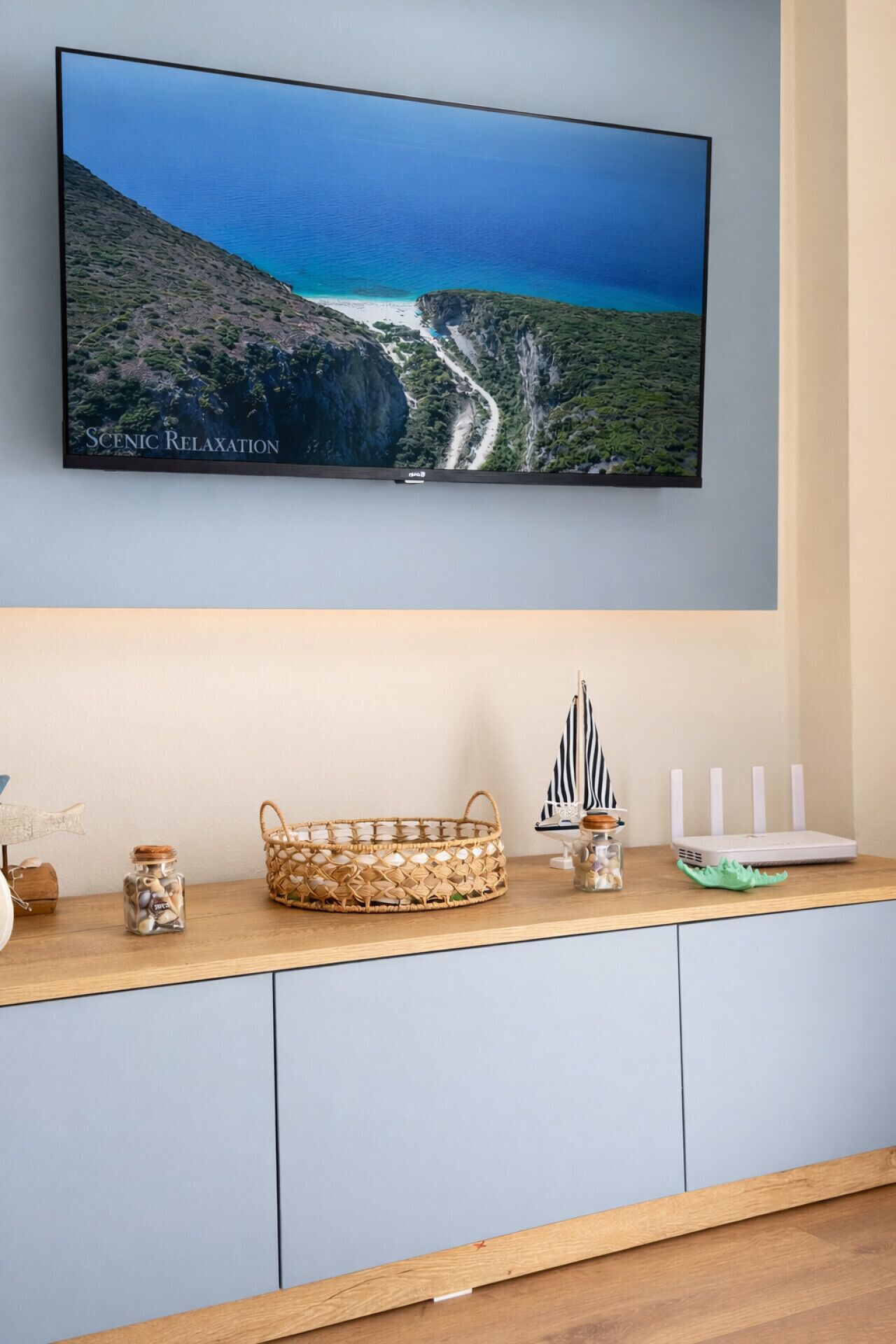 Un televisor de pantalla plana montado en una pared mostrando un paisaje costero. Debajo del televisor, un mueble de madera alberga una cesta de mimbre, un modelo de barco pequeño y un frasco de conchas marinas.