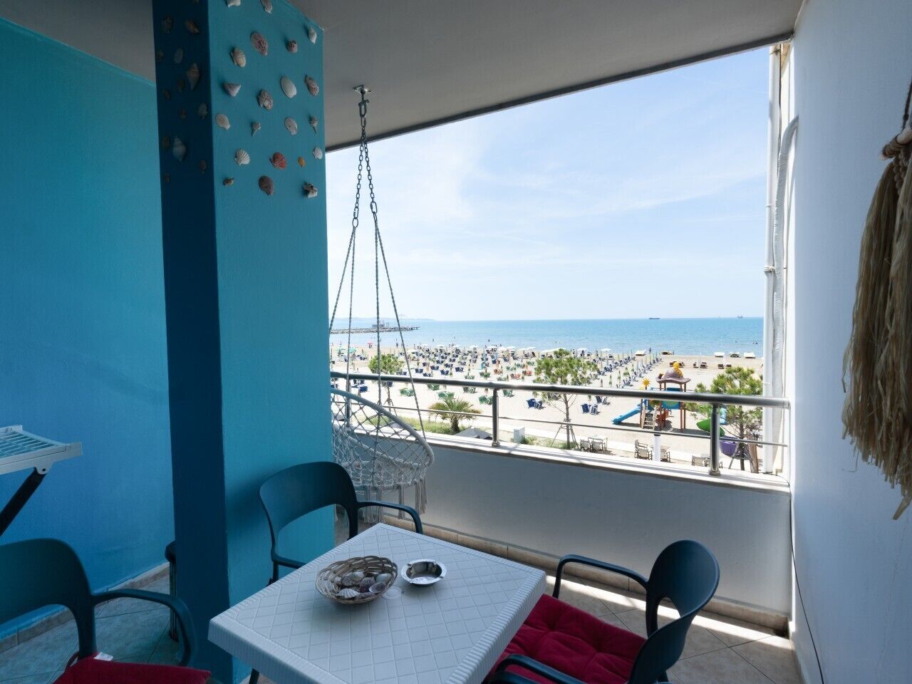 Varanda com mesa e cadeiras, vista para uma praia com pessoas e guarda-sóis.