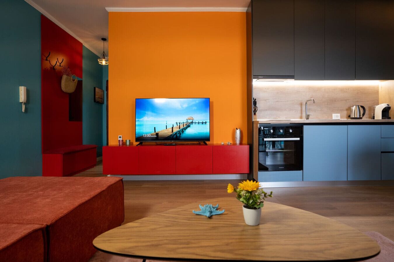 Un salon moderne avec un canapé rouge, une table basse en bois ornée d’une plante et un téléviseur diffusant une image de paysage.