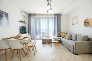 A modern living room with a grey sofa, a dining table with chairs, and a large window with sheer curtains. - Sea view-Sunny Apartment by TOK (Durrës)