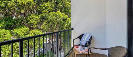 A balcony with a wicker chair, a small table, and a view of trees and a building.