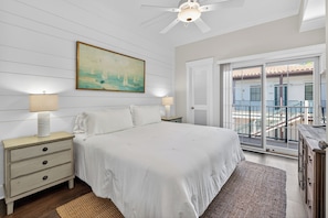 Une chambre à coucher avec un lit, deux tables de chevet avec des lampes, un ventilateur de plafond, un tableau et une vue sur un balcon doté d’une balustrade.