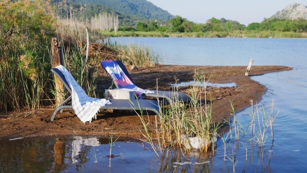 Un entorno junto al lago con una silla y una mesa cerca de la orilla, rodeado de vegetación natural y colinas al fondo.