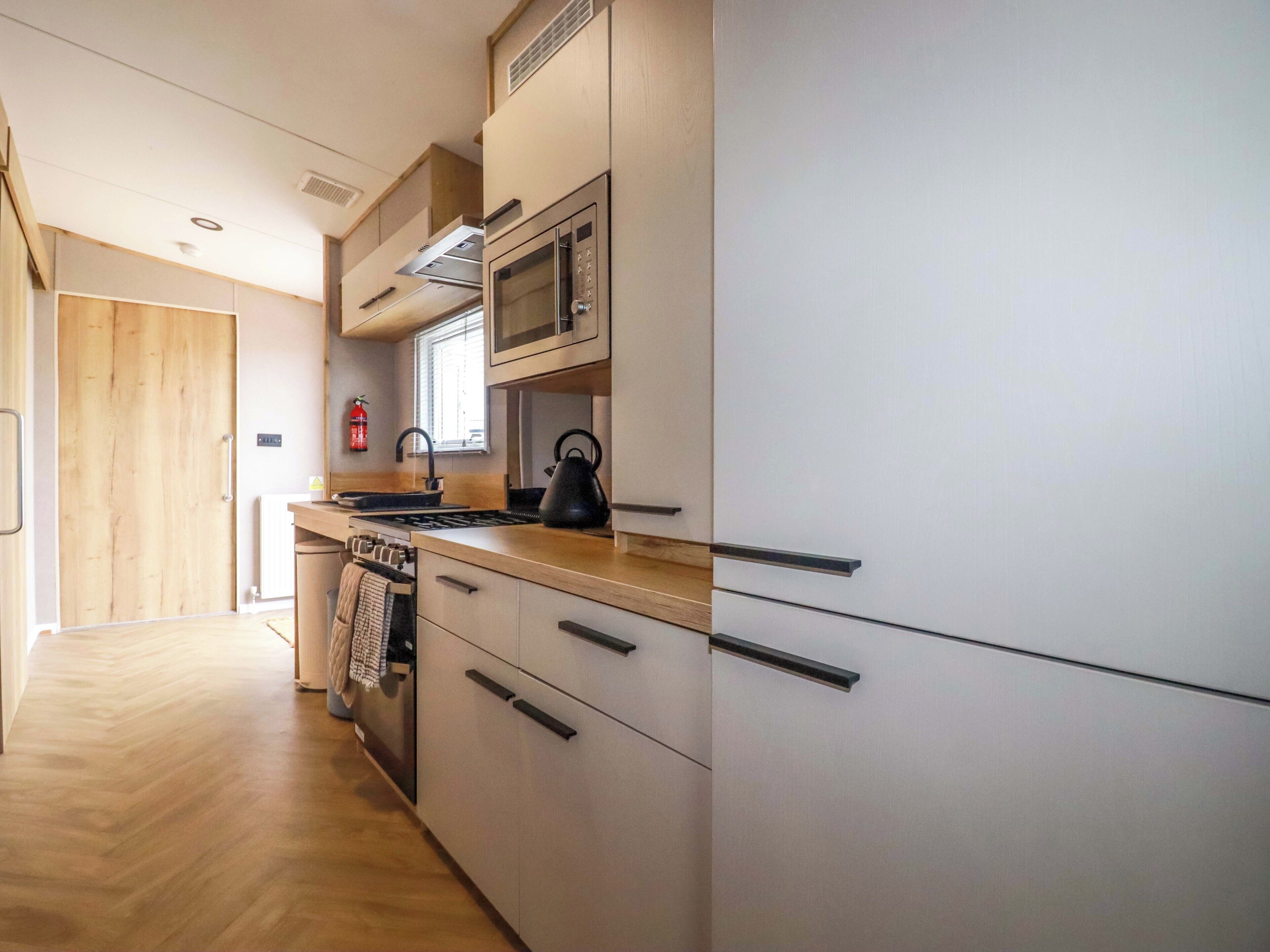 Una cocina moderna con microondas, hervidor y estufa. Se observa una puerta de madera y una ventana con persianas.