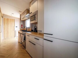 Una cocina moderna con microondas, hervidor y estufa. Se observa una puerta de madera y una ventana con persianas.