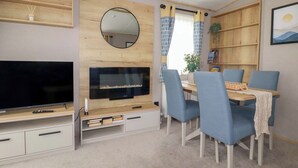 A compact living space with a flat-screen TV, a dining table with chairs, a bookshelf, and a mirror. - Coastal Escape (Par)