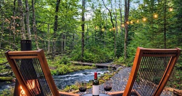 Zwei Korbstühle stehen einem Fluss in einem Wald gegenüber, dazwischen ein Tisch, eine Flasche und ein Weinglas.
