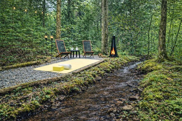 Waldlandschaft mit einer kleinen Außenbereich-Sitzgelegenheit, inklusive zwei Stühle, einem Tisch, einer Feuerstelle und einem Bach.