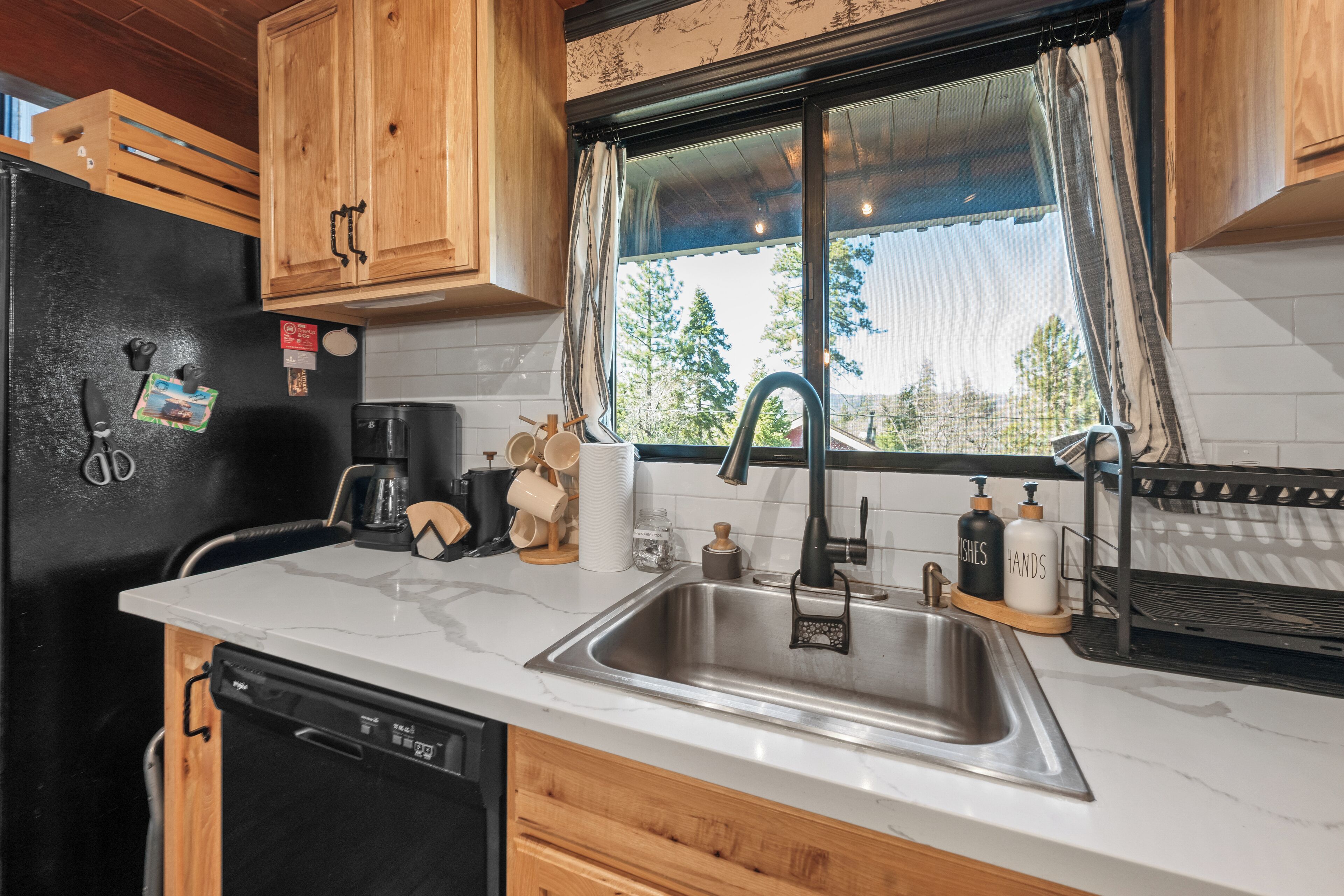 A kitchen with wooden cabinets, a black refrigerator, a stainless steel sink, and a window with a view of trees.