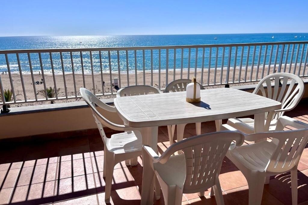 A terrace with a white table and chairs, overlooking a beach with a clear view of the sea.