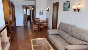 A living room with a sofa, a glass-top coffee table, a dining table with chairs, a wooden bookshelf, and a door leading to another room. - Spacious 4 bedroom apartment in first line of sea. Rodes Lux-61 (Calafell)