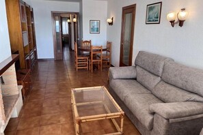 A living room with a sofa, a glass-top coffee table, a dining table with chairs, a wooden bookshelf, and a door leading to another room. - Spacious 4 bedroom apartment in first line of sea. Rodes Lux-61 (Calafell)