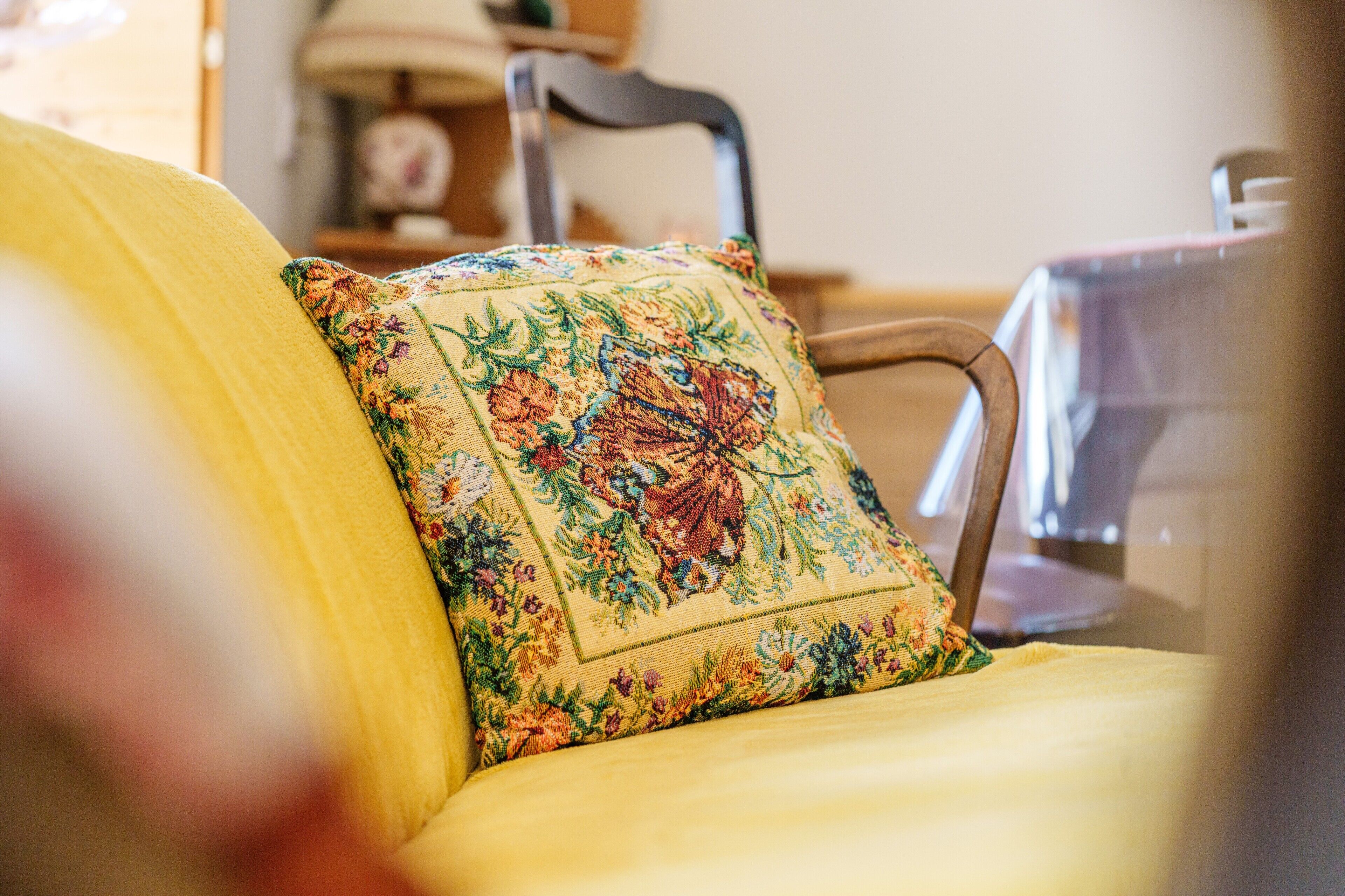 A yellow sofa with a floral pillow and a wooden chair in the background.