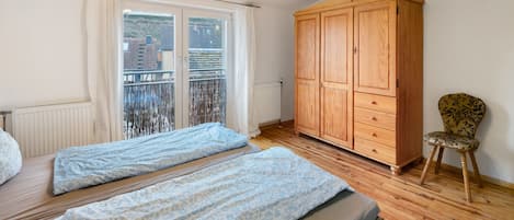 A bedroom with two beds, a wooden wardrobe, a chair, and a balcony with a view.