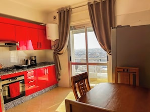 A kitchen with red cabinets, a gas stove, and a coffee maker. There is a dining table with chairs and a view of the city through large windows. - Stylish 2-bedroom apartment in Tangier with ocean views, central location, and beach access. (Tangier)